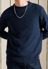 Pull en maille bleu marine avec un motif texturé, poignets et ourlet côtelés, et une petite écusson logo près de la ceinture. Accent chaîne argentée.