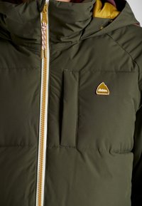 En närbild av en grön puffjacka med en gul inredning, som har en framzipper och en liten bergslogotyp på bröstet.