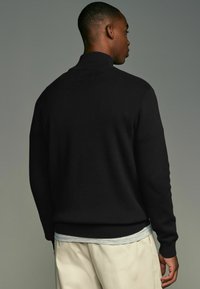 Schwarzer Strickpullover mit hohem Kragen, strukturierten Oberfläche und gerippten Bündchen. Über einem hellgrauen Hemd mit weicher, glatter Textur getragen.