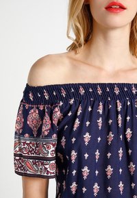 Haut marine à épaules dénudées avec encolure smockée, manches courtes à motifs, présentant des designs floraux et paisley en rose et blanc.