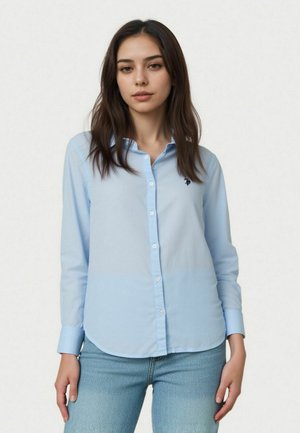 Giovane donna con capelli lunghi e scuri che indossa una camicia azzurra con bottoni e jeans blu, in piedi davanti a uno sfondo chiaro e uniforme.