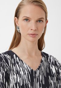 Schwarzes und weißes gemustertes Oberteil mit V-Ausschnitt, kombiniert mit übergroßen silbernen Ohrringen mit abstraktem Design.