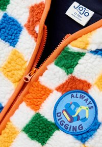 Fleece-Weste mit bunten Patchwork-Quadraten in Blau, Orange, Grün und Gelb. Front-Reißverschluss mit orangefarbener Einfassung. Verfügt über ein rundes Badge mit der Aufschrift "ALWAYS DIGGING."