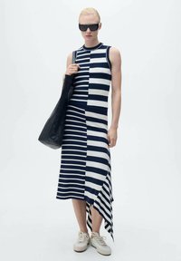 Geknipte mouwloze jurk in marineblauw en witte strepen, met een asymmetrische zoom, gecombineerd met een zwarte tas en lichte sneakers.