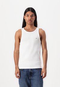 Weißes, geripptes Tanktop mit Rundhalsausschnitt, versehen mit einem kleinen Logo-Detail. Kombiniert mit blauen Jeans, die einen lässigen Schnitt zeigen.