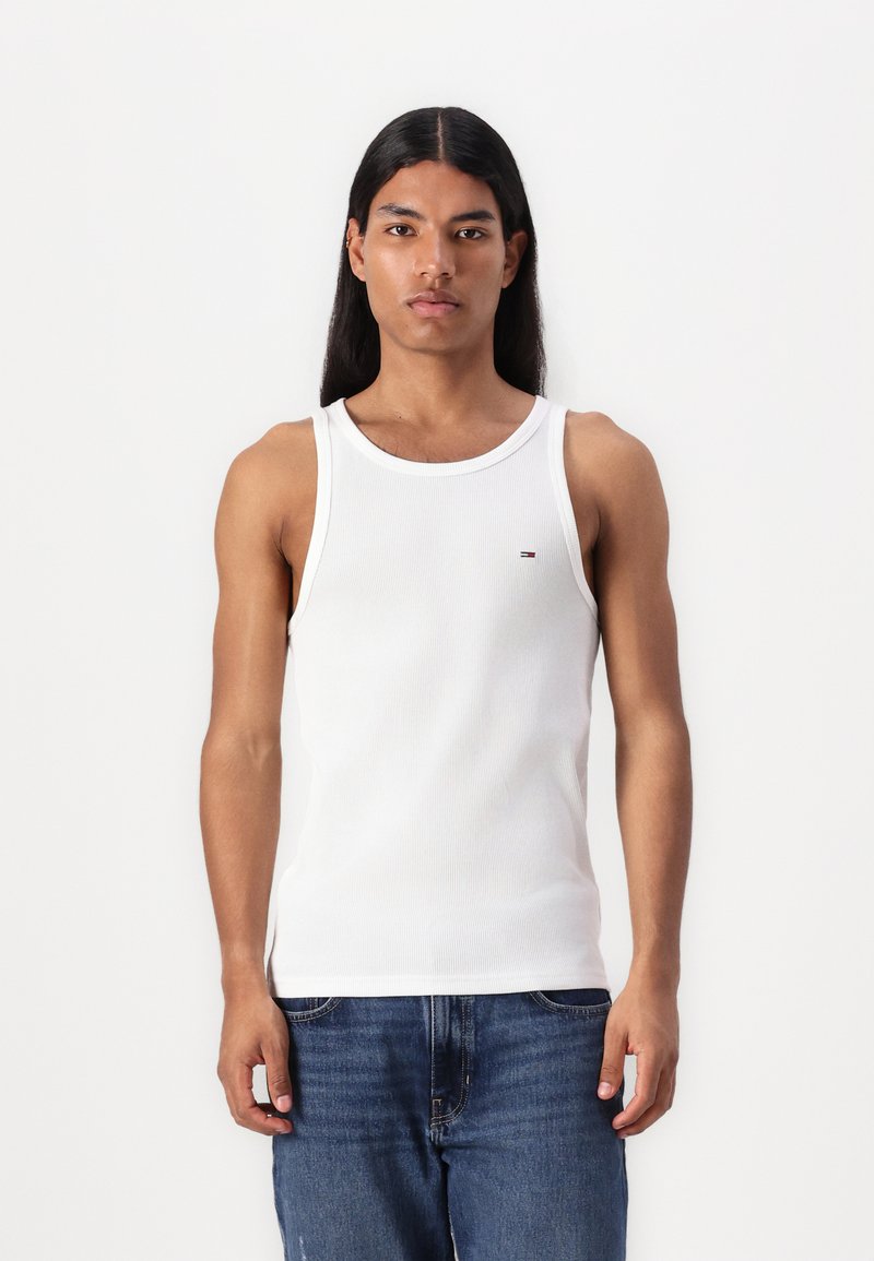Weißes, geripptes Tanktop mit Rundhalsausschnitt, versehen mit einem kleinen Logo-Detail. Kombiniert mit blauen Jeans, die einen lässigen Schnitt zeigen.