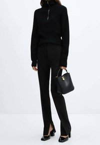 Pull en laine noir à col montant et demi-fermeture éclair, associé à un pantalon noir ajusté avec des fentes sur les côtés. Accessoirisé avec un petit sac à main noir.