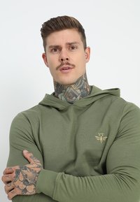 Un hoodie vert à coupe décontractée, doté d'une capuche avec cordon de serrage et d'un petit emblème doré sur la poitrine. Tissu lisse avec des manches longues.