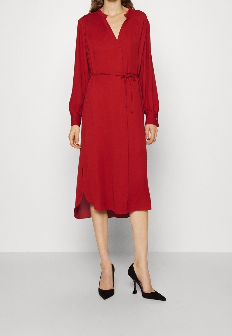 Rotes langärmliges Kleid mit V-Ausschnitt, tailliertem Bindegürtel und seitlichem Schlitz. Hergestellt aus glattem Stoff, mit entspanntem Schnitt und geknöpften Manschetten.