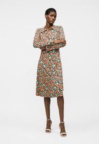 Femeie stând cu brațele încrucișate, purtând o rochie florală până la genunchi cu mâneci lungi și pantofi cu toc bej cu barete la glezne, pe fundal alb.