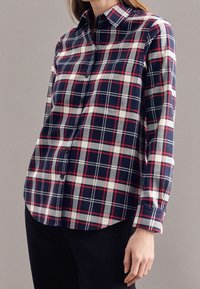 Red, white, and navy plaid button-up shirt with a collar, long sleeves, and a rounded hem, featuring classic plaid patterns and button closure.