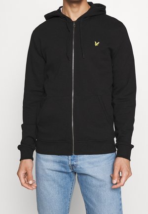 Mann trägt einen schwarzen Zip-Hoodie mit gelbem Vogel-Logo auf der Brust und hellblauen Jeans, steht vor einem einfarbigen Hintergrund.