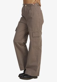 Cargo pants in taupe, made of durable fabric. Features a high waist, wide legs, side pockets, and a button closure. Textured, utilitarian design.