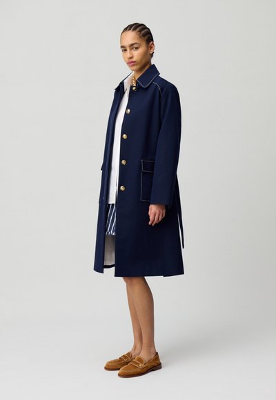 Manteau trench navy avec des boutons dorés, couture contrastée et poches latérales. Associé à une chemise blanche et une jupe rayée. Chaussures brunes à enfiler.