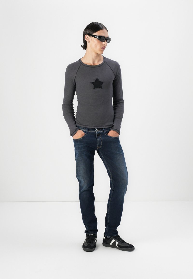 Graues Langarmshirt mit schwarzem Stern-Patch, kombiniert mit dunkelblauen Jeans und schwarzen Sneakers mit weißen Akzenten. Glatte Stofftextur.