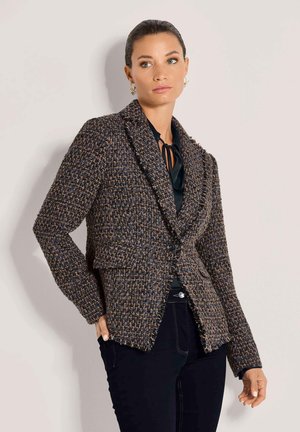 Frau trägt einen strukturierten braun-schwarz gemusterten Blazer über einer schwarzen Bluse und dunklen Hosen, steht vor einer schlichten hellen Wand.