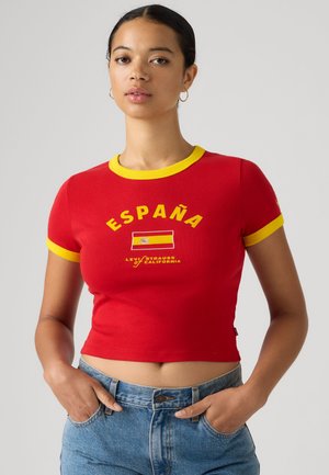 Mladá žena v červenom krátkom tričku so žltým lemom a textom España na španielskej vlajke, v kombinácii s modrými džínsami, ruky v vreckách.