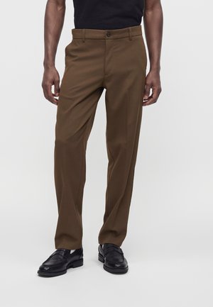 Man die bruine rechte broek, zwarte loafers en een zwart overhemd draagt, staand tegen een effen lichte achtergrond.