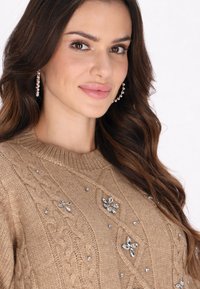 Beiger Strickpullover mit Kabelmuster, verziert mit floralen Motiven und Strasssteinen. Hat einen runden Ausschnitt und strukturierte Stoffqualität.