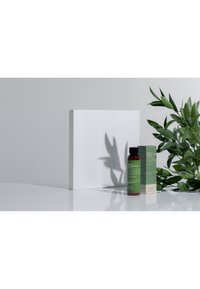 Röd flaska med lotion och svart kork, grön etikett bredvid matchande låda på en vit yta, omgiven av gröna blad och skuggor.