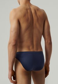 Męski model z wyrzeźbionymi plecami, ubrany w granatowe slipy, stojący z relaksującymi ramionami na jednolitym jasnoszarym tle.