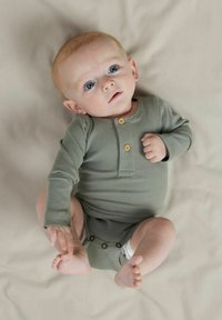 Sinine silmadega beebi, kes kannab rohelist soonikuid ülikonda, lamab beežil pinnal ja puutub vasaku käega oma parema jalaga.