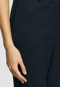 Pantalon noir avec un motif de petits points blancs subtils, texture lisse et poches latérales. La ceinture comporte une fermeture à bouton visible.