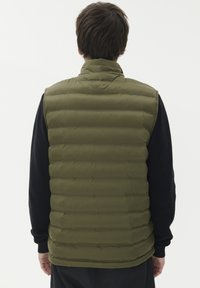 Moški v olivno zeleni prešiti brezrokavnik in črni dolg rokav, viden od zadaj proti enotni beli ozadju.