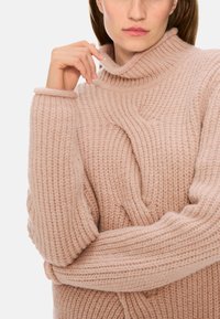 Helles rosa gestricktes Sweatshirt mit hohem Rollkragen und gerippter Textur; verfügt über übergroße Ärmel und ein verdrehtes Vorderteil.