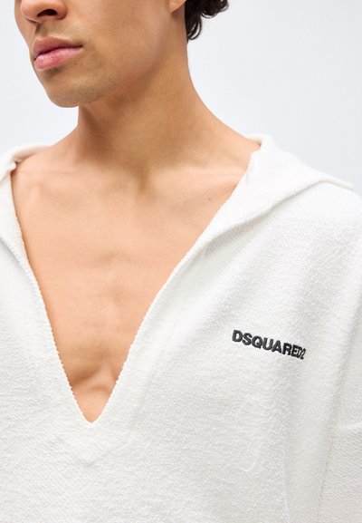 Άνδρας που φοράει ανοιχτό λευκό, υφασμάτινο hoodie με βαθύ V-λαιμόκοψη και το λογότυπο "DSQUARED2" στο αριστερό στήθος.
