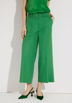 Femme portant un pantalon large taille haute vert avec fermeture à double bouton et des escarpins vert assortis à bride arrière, sur un fond uni.