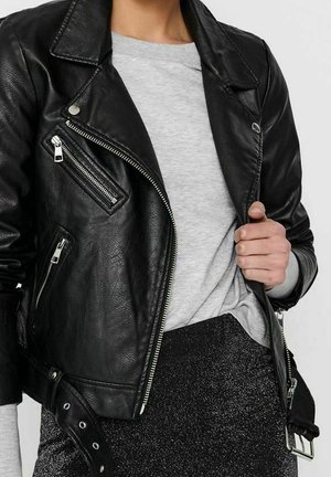 Personne portant une veste en cuir noire avec des détails zippés sur une chemise gris clair et un pantalon noir scintillant, tenant le col de la veste.