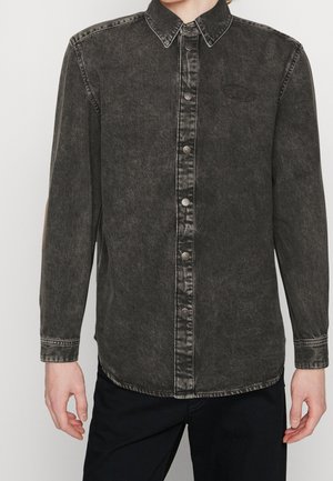 Camisa de mezclilla negra con cuello de botón, botones en la parte delantera y un sutil bordado del logo. Textura ligeramente desgastada y dobladillo curvado.