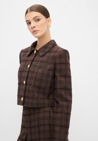 Blazer a scacchi marrone con design accorciato, colletto e bottoni dorati. Tessuto testurizzato con una leggera lucentezza e linee scure a contrasto.