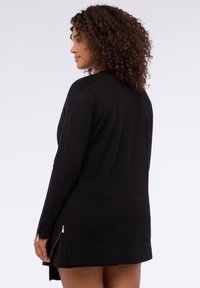 Schwarzer langer Cardigan mit gerippten Strickärmeln, seitlichen Reißverschlüssen, geradem Schnitt und mittellanger Länge, aus einem weichen Stoff gefertigt.