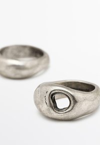 Anelli di metallo argento con una forma organica e liscia. Uno presenta un ritaglio ovale centrale; l'altro è un semplice band arrotondato con una finitura testurizzata.