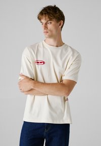 Cremefarvet t-shirt med korte ærmer, featuring røde logoaccents på brystet og ærmet. Lavet af blødt bomuldsstof.