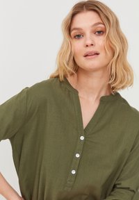 Blouse légère vert olive avec une finition texturée, dotée d'un décolleté henley à boutons et de trois boutons à l'avant. Manches courtes.
