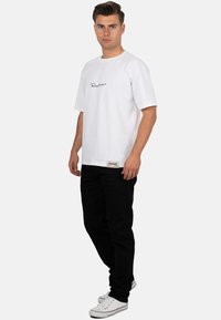 T-shirt bianca in cotone con logo ricamato nero, collo rotondo e maniche corte, abbinata a pantaloni neri e sneakers bianche.