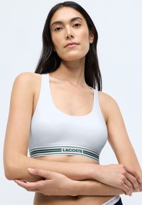 Balta kokvilnas bralette ar platu zaļo svītru jostu, kurā redzams "LACOSTE" logos, izstrādāta ar apaļu dekoltē un bez polsterējuma.