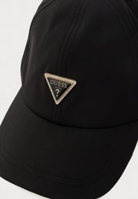 Cappello nero realizzato in tessuto liscio, caratterizzato da una toppa triangolare con il logo GUESS decorata con strass. Dettagli cuciti lungo la corona.