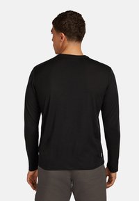 Man met kort, krullend haar draagt een zwart overhemd met lange mouwen en grijze broek, staand met zijn rug naar de camera tegen een effen lichte achtergrond.