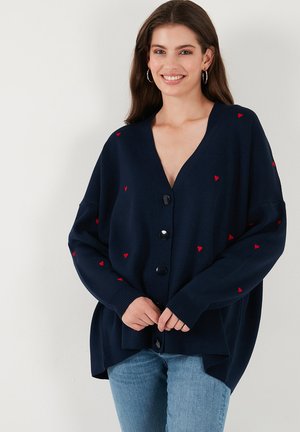 Femme portant un cardigan oversized bleu marine avec des motifs de cœurs rouges et de gros boutons, associé à un jean bleu clair, souriante contre un mur blanc.