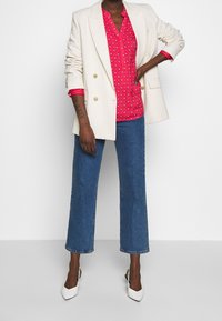 Blazer bianco doppiopetto sopra una blusa rosa a fantasia con scollatura a V, abbinato a jeans a zampa d'elefante blu e décolleté bianchi a punta.