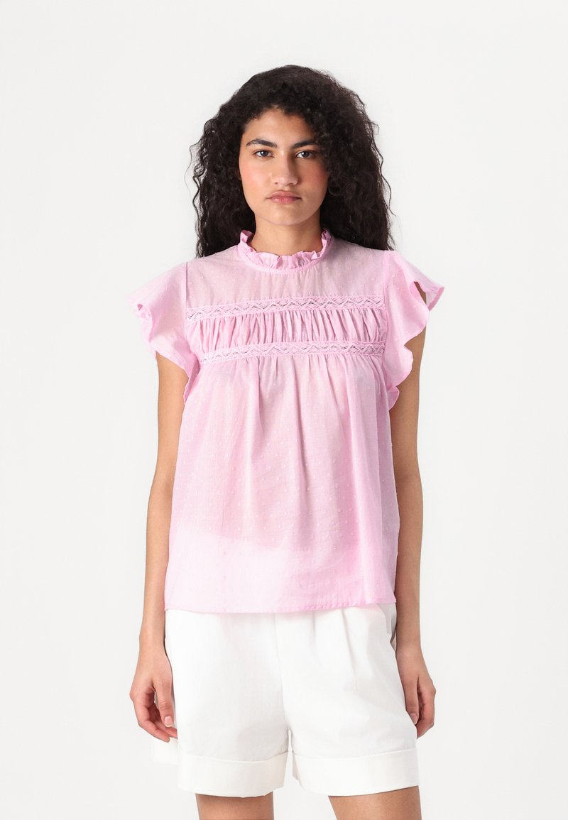 Blouse rose à manches courtes avec un col volanté, texture douce et motif à pois subtil; présente un tissu froncé au niveau de la poitrine.
