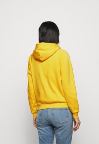 Gul hoodie med dragsko i huvan, ribbade ärmar och en känguruficka, parat med blå jeans. Slät textur med enfärgad design.