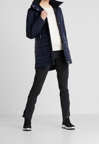 Marinblå quiltad jacka med huva, kontrasterande svart innerfoder, sidoreglar och en slät textur, kombinerad med svarta byxor och ankelstövlar.