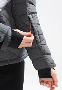 Grå quiltad jacka med en texturerad yta, med en orange dragkedja på ärmen och svarta innermuddar.