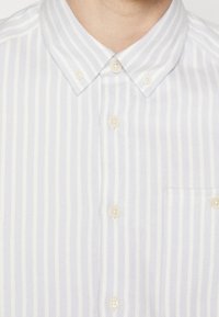 Vaaleansinisävyinen ja valkoraidallinen nappipaita, jossa on kaulus, helmiäiskoristeiset napit ja rintatasku. Kangas näyttää hienovaraiselta tekstuurilta.