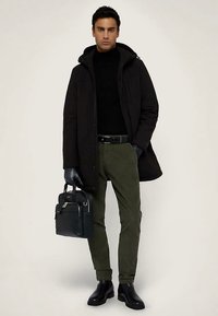 Černá parka, černý rolák, olivově zelené kalhoty, černé kožené rukavice, černé kožené boty. Drží tmavou tašku se stříbrným akcentem.